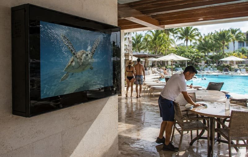 Custodia per TV da esterno in policarbonato nero che protegge una TV da 65 pollici accanto alla piscina di un hotel dagli schizzi di umidità e dal traffico degli ospiti